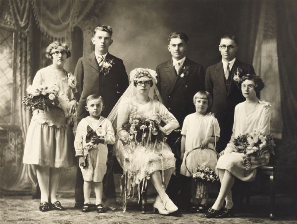 Group portrait in front of a painted background of a wedding party. Three men and three women pose with a young girl and boy. The three women and one of the men are wearing round eyeglasses. Persons unidentified.