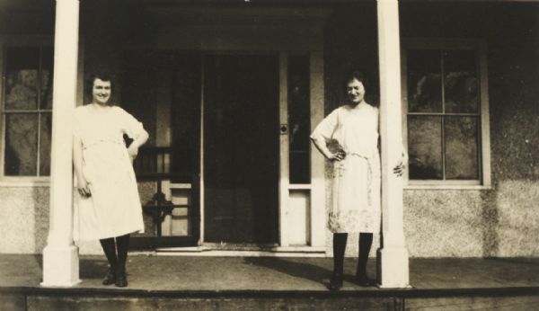 Two young women posing while leaning on the columns of a front porch.