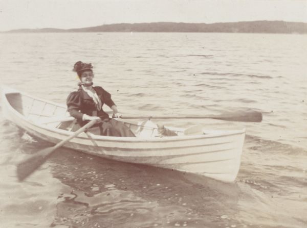 A woman wearing a hat, high-collared blouse, jacket, and long skirt sits in a rowboat while rowing a small boat, presumably on Lake Mendota. A cat sits at the bow of the boat. Inscribed on back of original mount: "Prof. Mrs. Daniels."