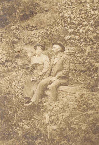 Man and woman seated on an overgrown hillside. The woman has ferns on her lap and is holding an umbrella. Inscribed on reverse of original mount: "B.D.L. and P.B. Dudley at Spring Brook, Delton, Wisconsin."