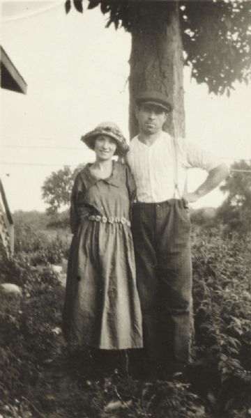 Farm couple standing under a tree. The roof and stair railing of a building is on the left.