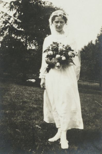 Full length portrait of a bride in a long white dress and wearing a veil standing on a lawn holding a bouquet of flowers. There are trees in the background.