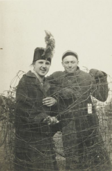 Portrait of a man and a woman posed together, wrapped in wire fencing. It must be a prank because they are both smiling. She is wearing a hat with a plume on the front, and a coat. He is wearing a jacket and cap and appears to have a pack on his back.