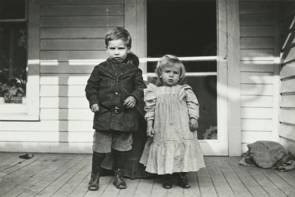 Two children, a boy and a girl, stand on a porch. A woman in a dress (face obscured) crouches behind the two children.
