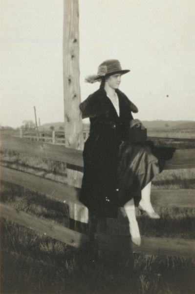 A fashionably dressed young woman is posed sitting on a fence. She is wearing a long coat over a dress, a hat with a wide brim, and high-button boots. Her hands are folded over a handbag in her lap.
