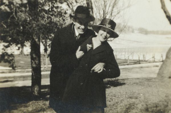 A young couple wearing coats and hats are posing outdoors.