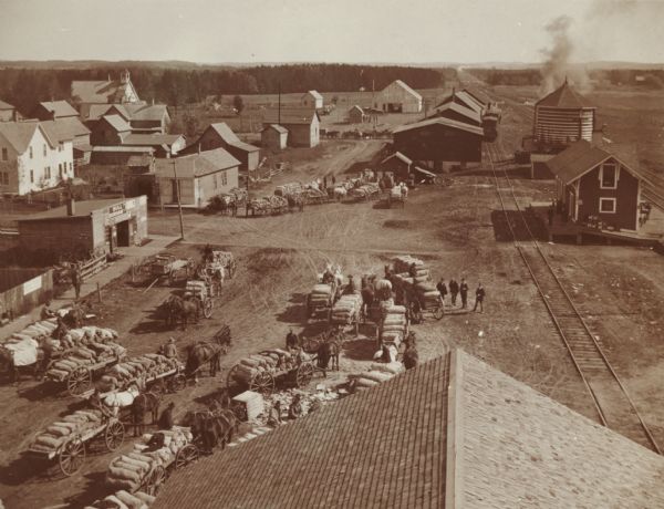 Elevated view of farmers with horse-drawn wagons waiting to ship their harvest goods, probably potatoes, at an unidentified town. There are railroad tracks, a depot and a water tower on the right.