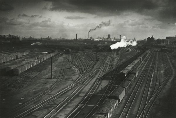 Elevated view of railroad yards with trains sitting on many of the tracks, with other tracks sitting empty. Steam is rising from trains in the background, as well as from smokestacks in the distance.