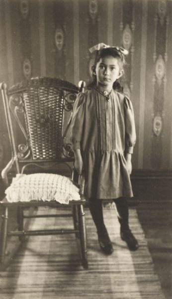 Portrait of a young girl standing beside a wicker-back chair.