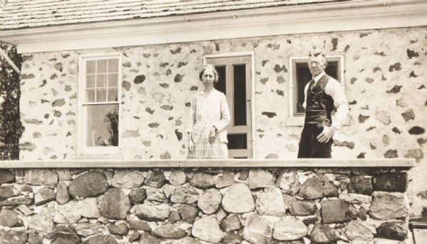 Mr. and Mrs. Charles D. Stewart standing on the porch of their home. The dwelling and porch railing are built of stone and mortar.