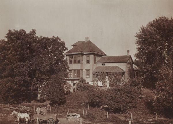 Farm home of Thomas Convey family, at or near Ridgeway. In the foreground, two men stand near a horse-drawn wagon. Five women are standing on a stone wall near the porch of the house, which is on a hill. A woman stands on the porch which is covered with vines, and a man stands in the open doorway of a shed or garage on the left.