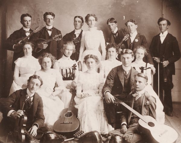 Group portrait of the Brodhead Mandolin Club. The men are wearing suits and neckties, and all the women wear long white dresses. There are mandolins, as well as a violin and a guitar.