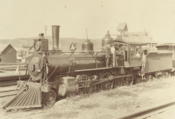 Wisconsin Central railroad locomotive no. 120 at the railroad station. Engineer Clelland McVick is standing in the gangway, and a woman who is presumably his wife is seen at the cab window. Two other men and another woman are also posed on the locomotive.