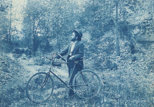 Cyanotype print of a man standing and holding a bicycle in a wooded landscape. Probably a self-portrait of William H. Dudley of Madison.