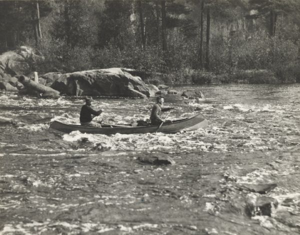 View from shoreline of two men in a canoe running the Cedar Rapids on the Flambeau River.