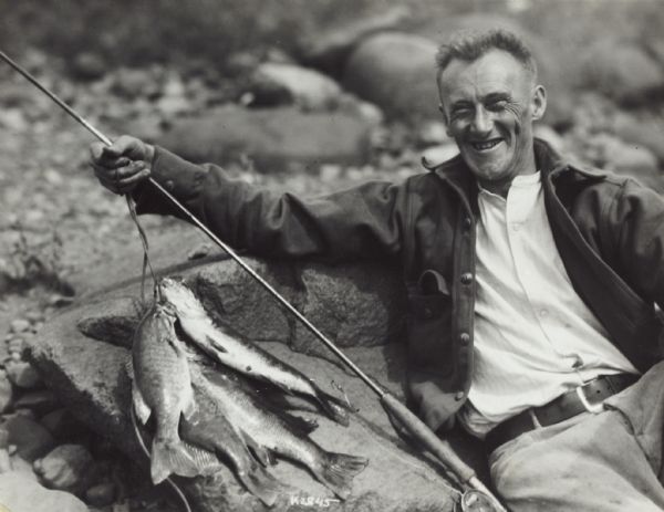A fisherman poses on rocks with his fishing pole and a stringer of fish.