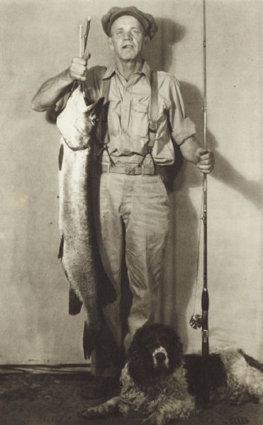 A man wearing suspenders and a hat stands holding his catch of a large fish. He holds his fishing pole in his left hand, and a dog is lying at his feet.