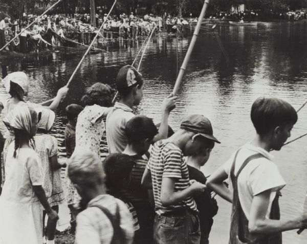 View of large crowd of boys and girls fishing along the shoreline with bamboo poles at a lake. Very likely a sponsored "fishing contest." There are a few people out on the lake in boats.