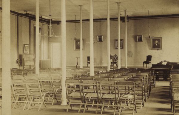 View from back of room of the interior of the chapel at Rockford College. There is a piano on the right, in front of the stage. Folding chairs are set up in rows between the columns in the room. The original land for this college was purchased from Buell G. Wheeler. William Lathrop was chairman of the executive committee in 1891.