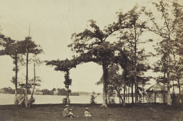 Lakeside lawn set up for croquet game and garden pavilion at the estate, "Sunnybank." A man sits in the grass with two children. On the right in the background is a pavilion among trees. Across the lake are houses and buildings.