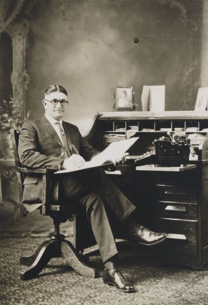 Unidentified business or professional man seated in a chair in front of a roll-top desk. He is wearing round eyeglasses, and there is a typewriter on the desk. In the background is a painted backdrop.