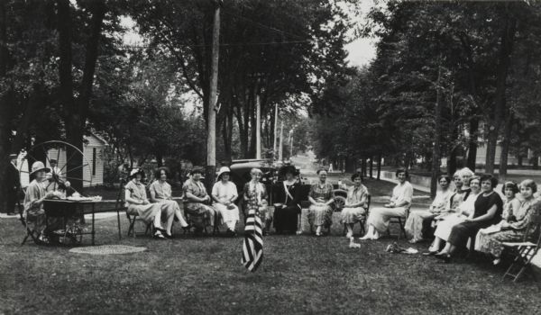 Group of ladies at an outdoor tea during "homecoming." Behind the woman on the left is a large spinning wheel. An American Flag is standing on the lawn in front of the group. Two men stand in the background on the left.