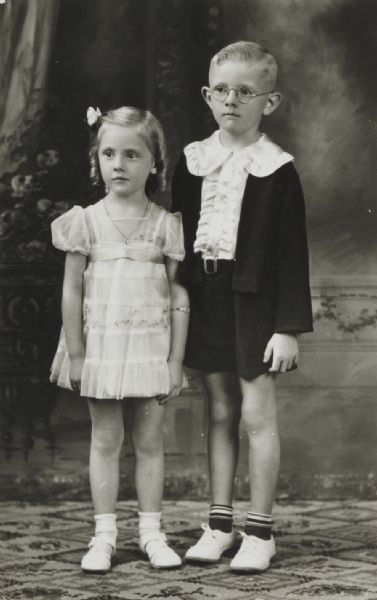 Full-length studio portrait in front of a painted backdrop of a boy and girl, probably brother and sister.