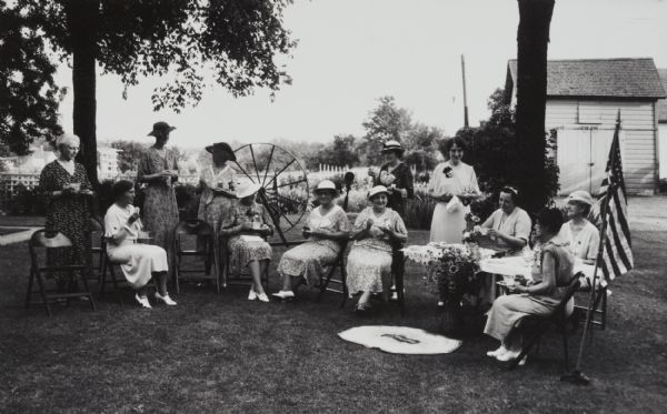 Group of ladies sitting and standing at an outdoor tea party during "homecoming." There is a large spinning wheel on the lawn behind the group.