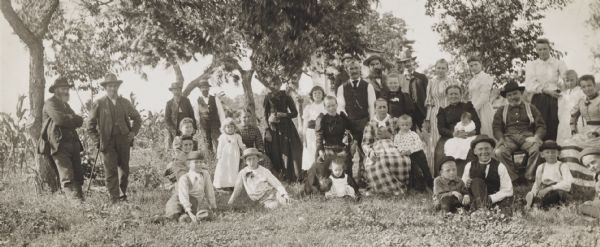 Large group of farm adults and children, probably of German extraction, of one or more families, gathered under fruit trees at the edge of a cornfield, presumably at or near Alma.