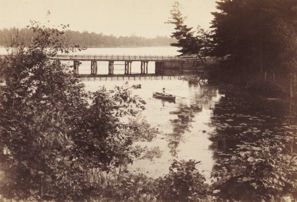 View from shoreline looking down on Mud Creek, about two miles above De Pere, showing small bridge and a man in a rowboat.