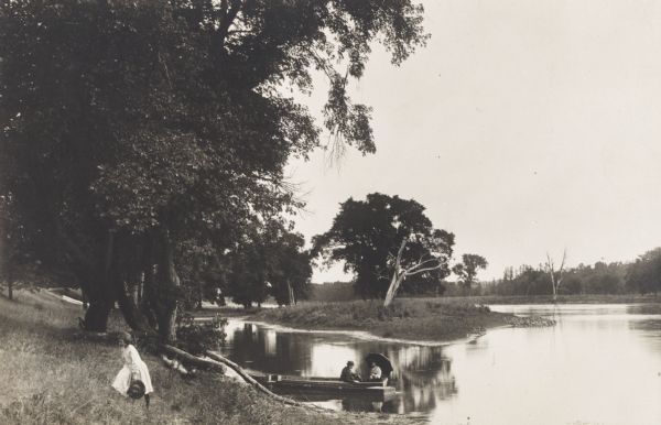 View along bank of river towards a young girl walking in the grass near the shoreline. Behind her, a man and woman sit in a rowboat on the Rock River. They are about one mile below Oregon, Illinois.
