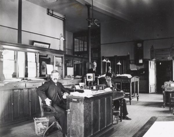 Interior of the Knapp, Stout & Co. lumber mill office with posed members of the staff. On the desk in the center of the room is a device with an earpiece hanging on the side of a box with a speaker tube. The box is attached at the back to a curved pipe. It is either a wall telephone, or an intercom.