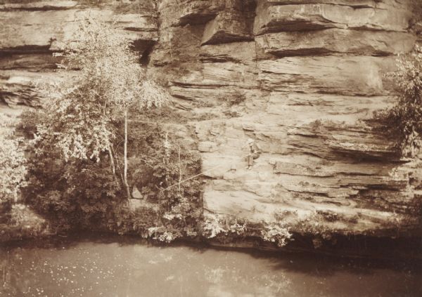 Elevated view over water towards an elderly man using a long pole to fish from a steep rock ledge on the opposite shoreline.