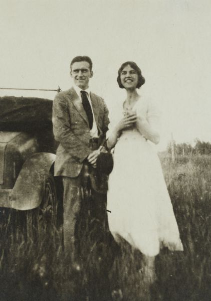 Unidentified smiling couple standing in a field near an automobile.