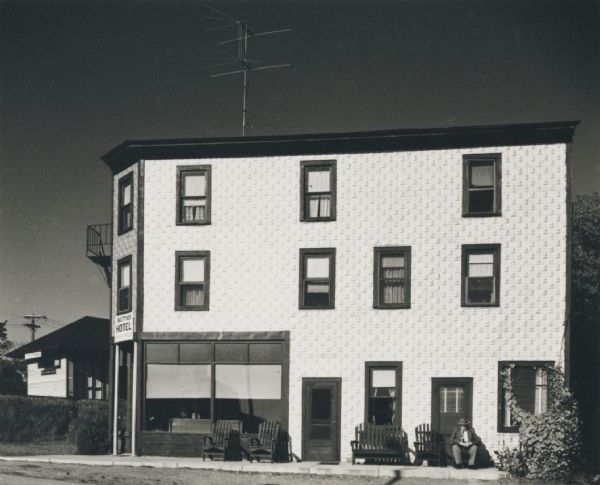 Exterior of a hotel, with bright metal siding in full sunlight. An elderly man is seated in a chair outside. Behind the hotel on the left is what appears to be the Gresham train station.