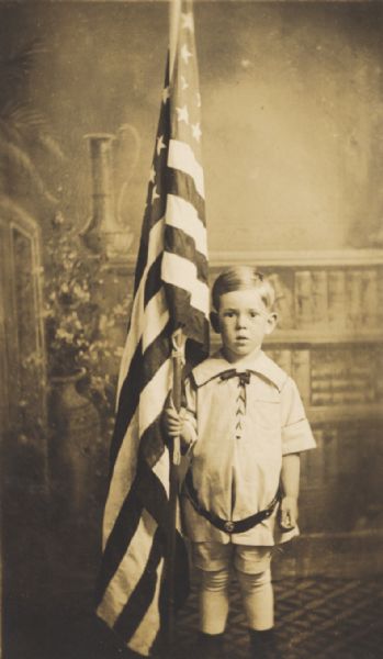 Studio portrait in front of a painted backdrop of a small boy holding a large U.S. flag.