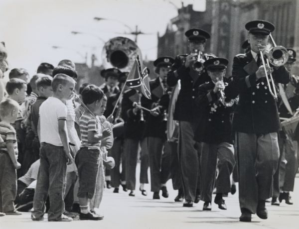 View along side of street towards a teenage brass band marching down the street in a Memorial Day parade in West Allis. A group of young children stand on the left watching the parade.