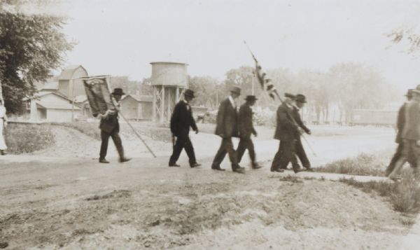 Small group of elderly men, two of them bearing, respectively, a U.S. Flag, and a banner, perhaps a company roster. This may be a group of Civil War veterans marching on Memorial day. Behind them are industrial buildings, railroad cars and a water tower.