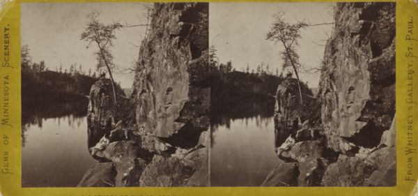Stereograph of the dalles of the St. Croix river, on the Wisconsin-Minnesota border. Along the shoreline on the right, a woman in a long dress is posing on a low rock, and a man wearing a hat is behind her posing on top of a steep rock.