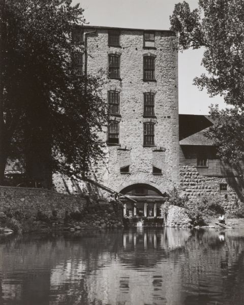 View across water of the Old Mill surrounded by trees and water. Water is coming out of an opening in an archway of the mill. On the left automobiles are parked, and a group of people, including some children, are sitting on top of the stone wall at the shoreline. On the right a man is squatting near a tree limb at the shoreline, perhaps fishing.
