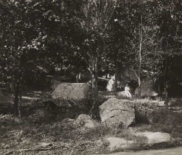 View towards hillside with an outcropping of large talus rocks surrounded by trees near Castle Rock village. In the foreground is what appears to be a dirt path, and partway up the hill is a barbed wire fence. A man wearing a hat and long white jacket with his arms folded is leaning against one of the rocks on the right just behind the fence.