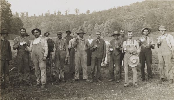Group of farm workers, men and women, posed in a field, holding various beverages. One man has his arm over a barrel on a stand in the center. In the background is a tree-lined ridge.