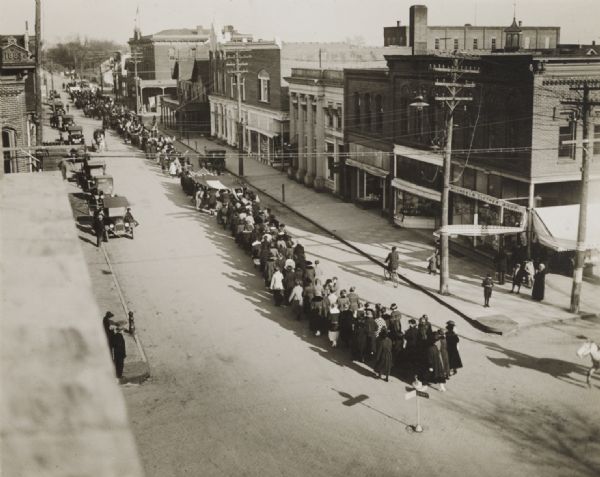 Elevated view from building of informal Armistice Day street parade at an intersection of the downtown area. They are celebrating the conclusion of the first World War. A large group of women are walking away from the intersection behind another group of women holding onto the sides of large flags. Pedestrians watch from the streets and sidewalks.