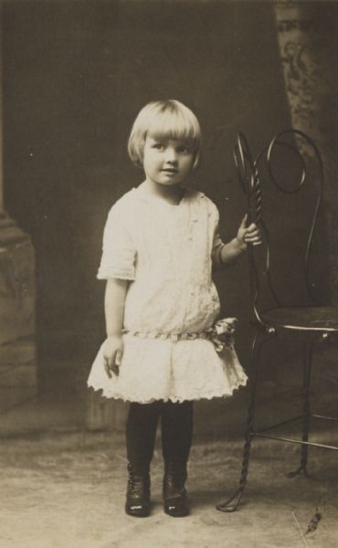 Full-length studio portrait in front of a painted backdrop of a small girl standing and holding on to a wrought-iron chair.