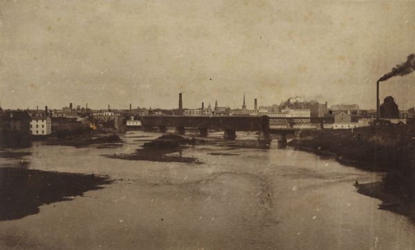 View down Rock River towards a covered bridge near another bridge, with factories and the city in the background.
