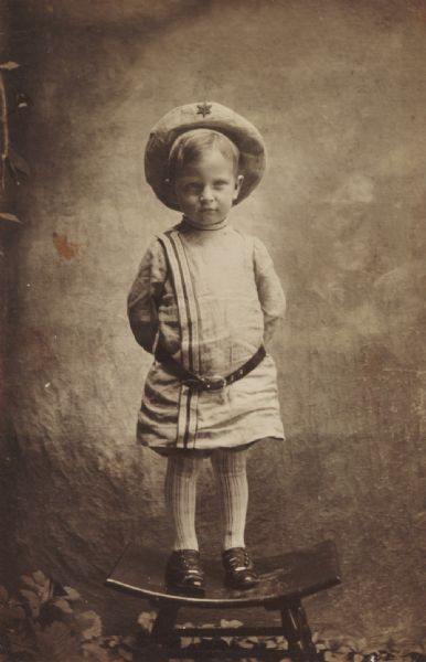 Full-length studio portrait of a young boy standing on a stool with his hands behind his back. He is wearing a tunic, or Buster Brown suit, striped stockings, button-up shoes, and a hat with a star on the front. Behind him is a studio backdrop and there are leaves or plants on the floor behind the stool.