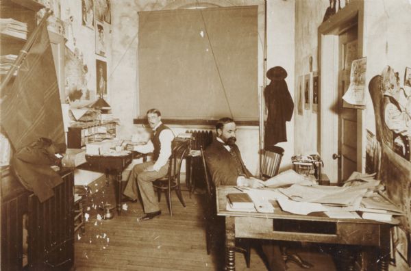 Two men sitting at desks in the editorial room of the <i>Milwaukee Journal</i>. The room was decorated by a newspaper staff artist. At left, the balcony scene from Romeo and Juliet; at right, a "Katzenjammer," a seated man with a cat beside him and his foot, swollen with gout, resting on a stool while a monkey (above the door) tickles his head with a head of barley. The room was eventually destroyed by fire.