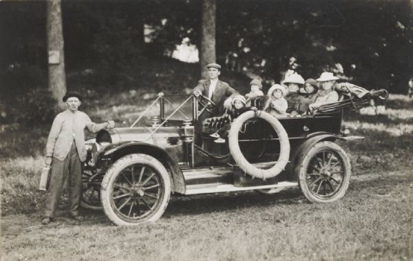 Family posing in an open touring automobile parked on a country road. An older man is standing at the front of the car holding a metal can.