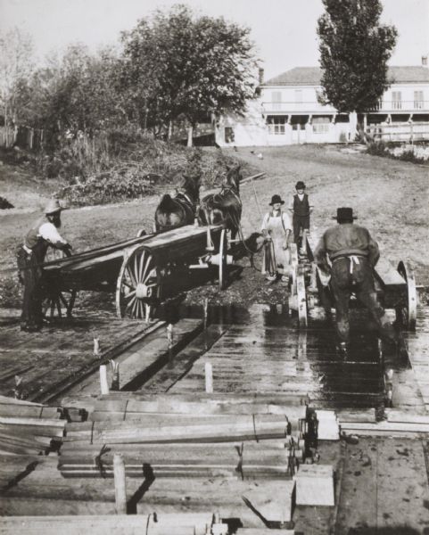 A raft of lumber which has been floated down the Wisconsin River to be broken up and sold. Two men are standing on the raft, each near lumber stacked on a cart, one driven by a team of two horses. Two other men are standing on the shoreline. Behind them a lawn slopes up towards a large building.