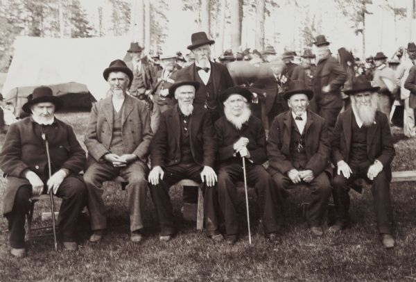 Seated, posed group portrait at the annual meeting, outdoors at the semi-centennial celebration in Sherman's Grove.  Identified from left: John Eckles, Ed. Metcalf, Jim O'Brien, Leander Trudell, Antervine Precout, and G.W. Franklins. Standing is S. A. Sherman. Behind them is a crowd standing among the trees near a tent.